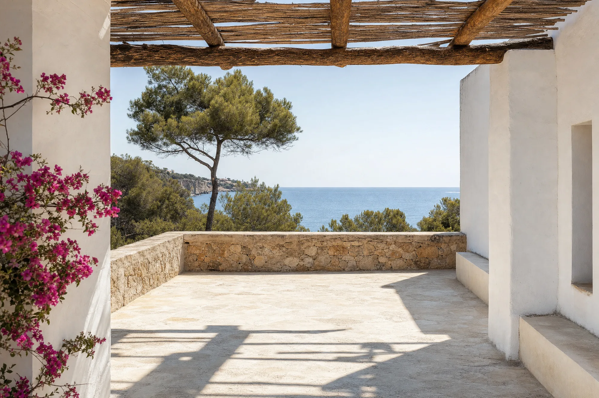 A whitewashed Balearic terrace under a timber pergola, looking out to the sea through umbrella pines
