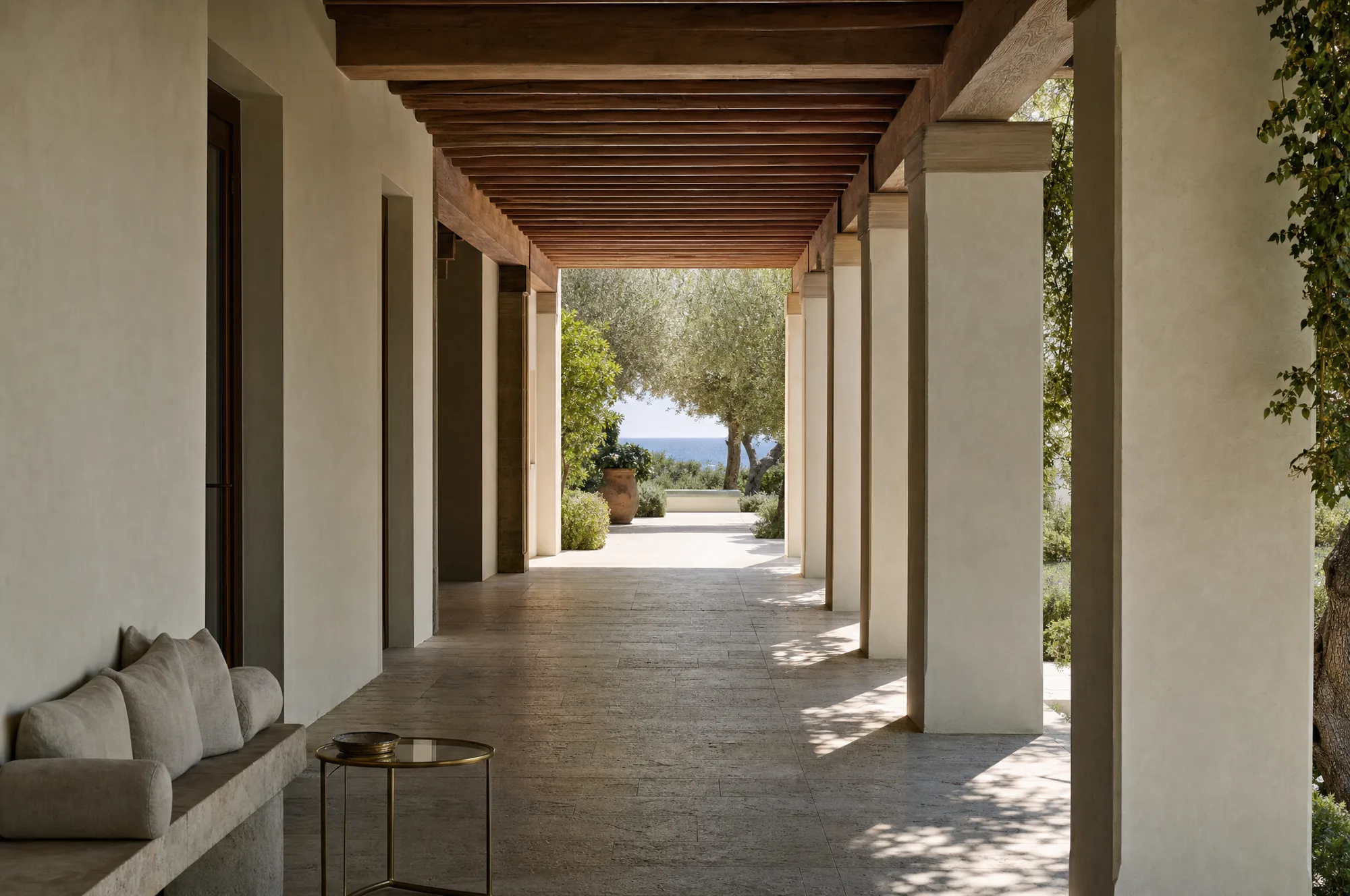 A deep shaded loggia of lime-rendered piers under a chestnut beam ceiling, bands of shade falling across a limestone floor with the bright garden beyond