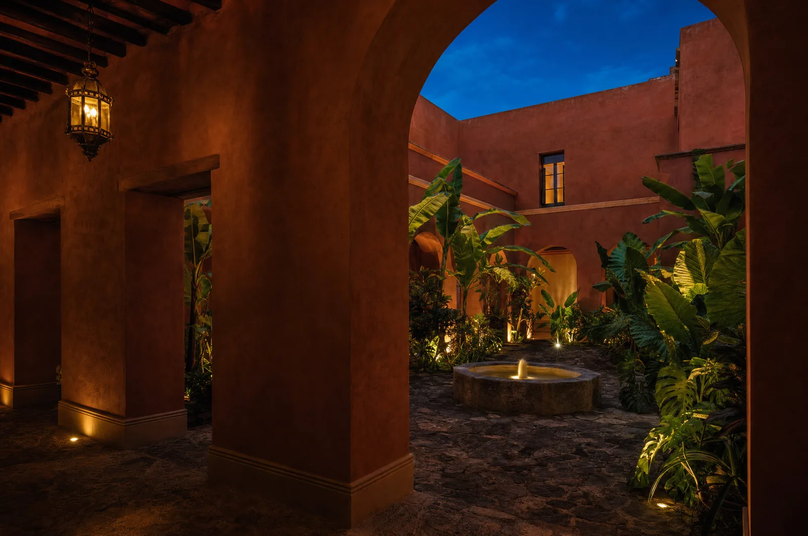 Patio interior de un hotel boutique mexicano al anochecer, muros de cal terracota, fuente de piedra y follaje iluminado desde abajo