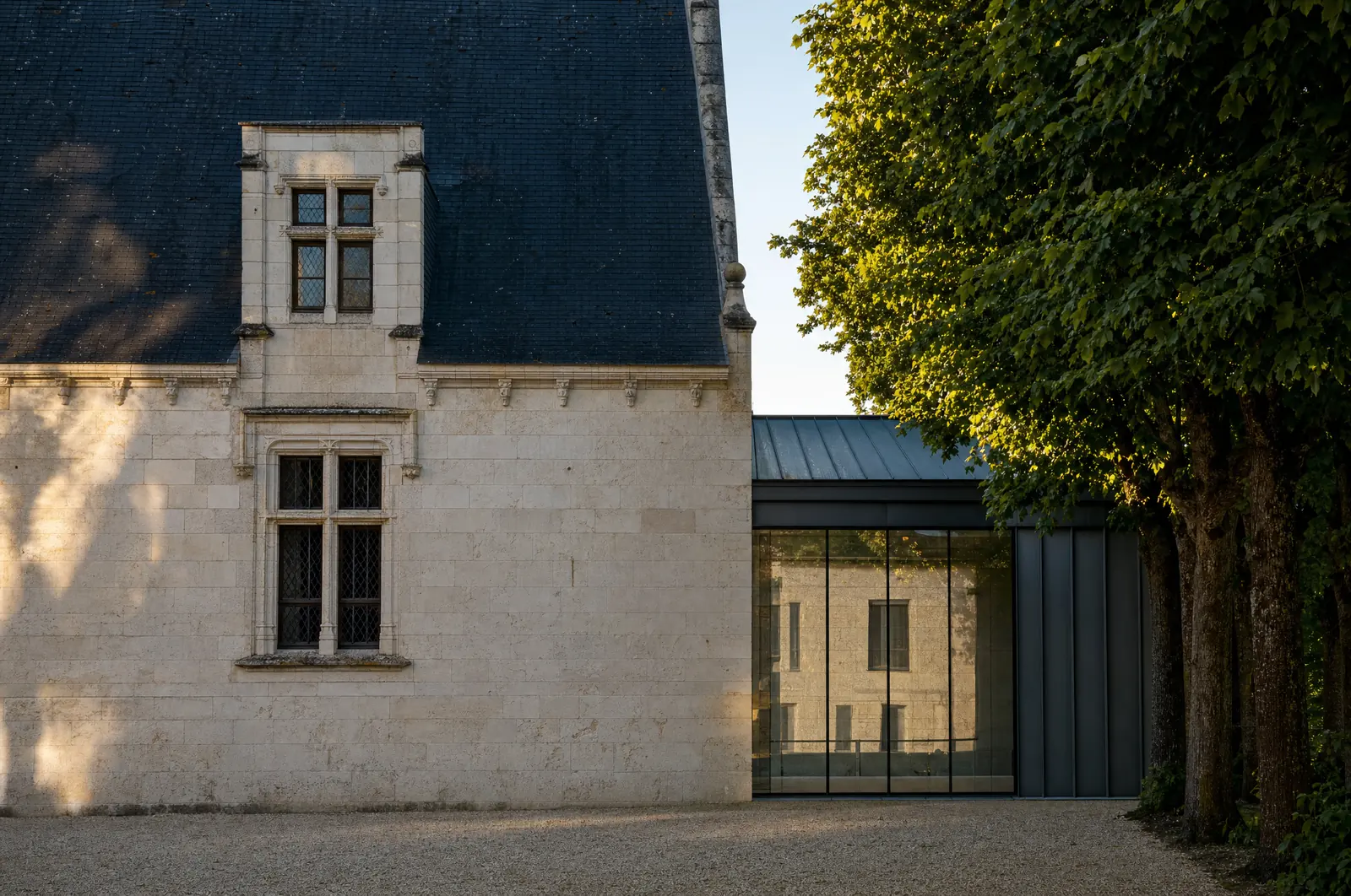 Pignon de tuffeau a fenetres a meneaux sous un toit d'ardoise, jouxtant une extension contemporaine de zinc et de verre qui reflete la facade ancienne